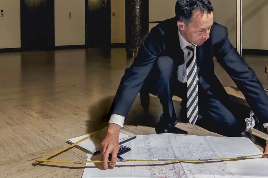 Mitarbeiter in Anzug kniet auf dem Boden und prüft ausgebreitete Baupläne sowie Vermessungsunterlagen zur Vorbereitung einer Bestandsaufnahme.