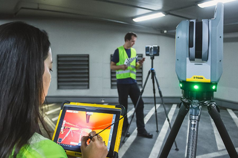 Mitarbeiterin bedient ein Tablet vor einem 3D-Laserscanner, während ein Kollege Vermessungsarbeiten in einem Gebäude durchführt.